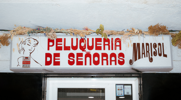 Storefront signage, white background At left: outline drawing of a woman's face. Text in red: "PELUQUERIA DE SEÑORAS." At right in brown and a different font, "MARISOL"